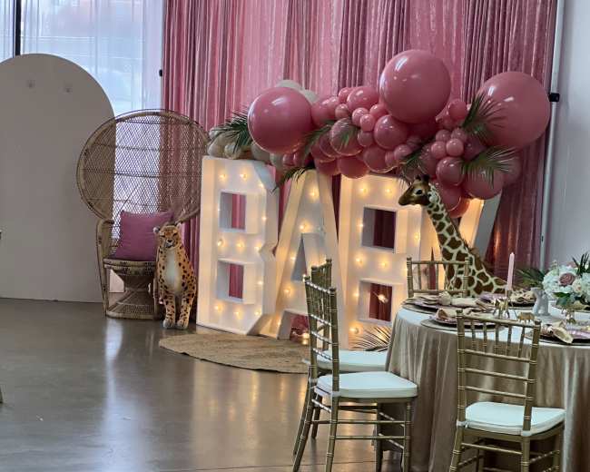 A decorated event space featuring a large illuminated "BABY" sign, a seated giraffe decoration, and a table set for a celebration.