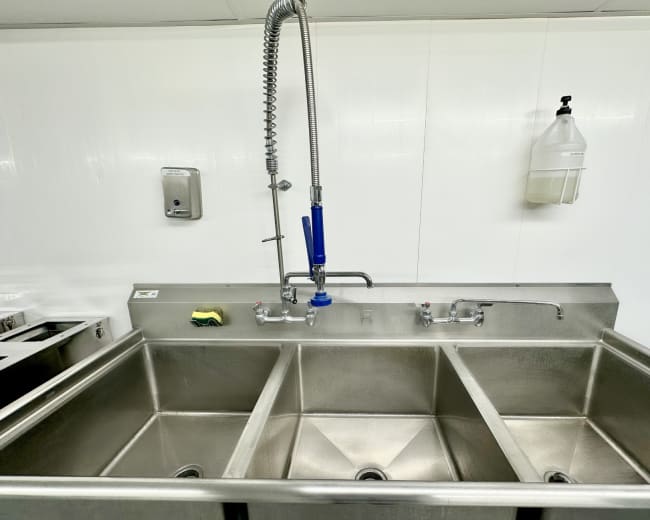 The image shows a stainless steel sink with three compartments, a faucet with a sprayer attachment, and a soap dispenser mounted on the wall.