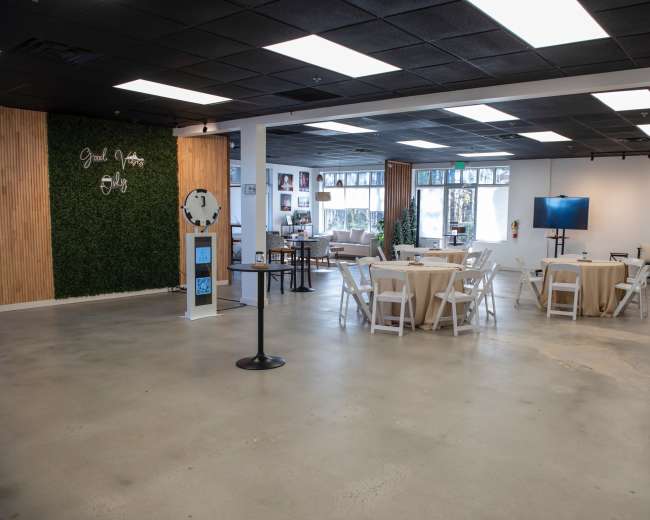 The image shows a spacious event venue with round tables dressed in beige tablecloths, a large screen, and a green vertical garden accent wall.
