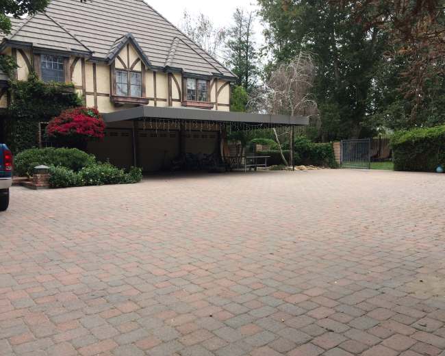A large brick-paved driveway leads to a house with a covered entryway adorned with greenery and flowers.