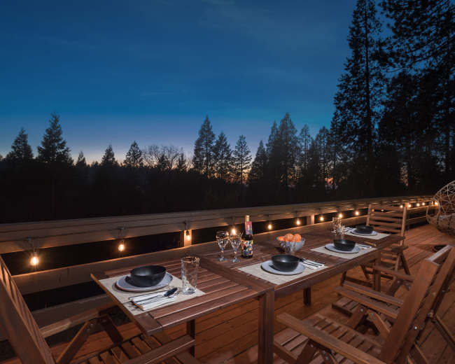 A wooden dining table is set with plates and glasses on a deck surrounded by tall trees under a twilight sky.