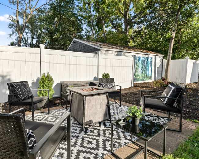 The image shows a outdoor seating area with black chairs and a fire pit, surrounded by white fences and greenery.