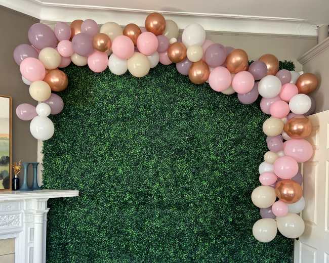 A decorated wall covered in artificial greenery, with a cluster of balloons in various colors arranged at the top.