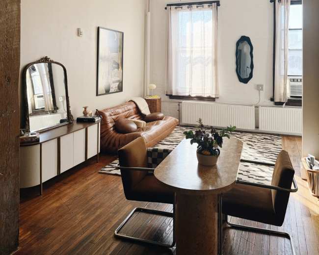 The image shows a minimalist living room featuring a brown leather sofa, an oval mirror, a dining table, and large windows with sheer curtains.