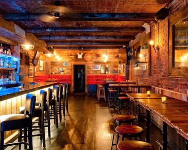 The image shows an empty bar interior featuring wooden walls, a long bar counter with stools, and dim lighting creating a cozy atmosphere.