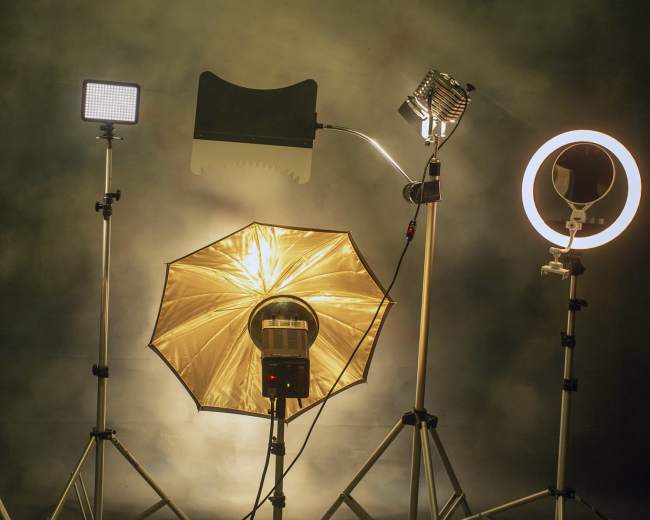 A collection of various photography lighting equipment is set up on tripod stands against a textured background.