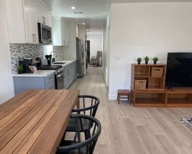 The image shows a modern kitchen and living area with a wooden dining table, black chairs, and a television unit against a light wall.