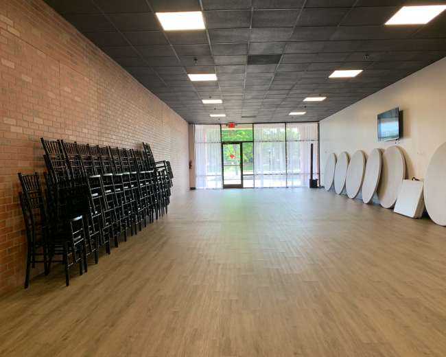 A spacious interior room with stacked chairs against a brick wall, round tables lined up along the opposite side, and large windows at the entrance.