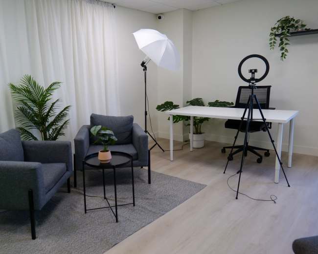 The image shows a well-lit, minimally furnished studio space with two gray chairs, a small table with a plant, a photography light setup, and a desk with a ring light.