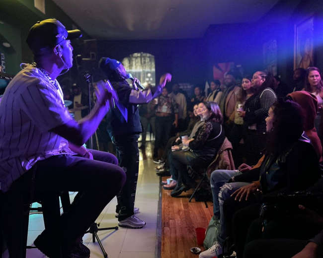 A performer sings on stage while an audience enjoys the show in a dimly lit venue.