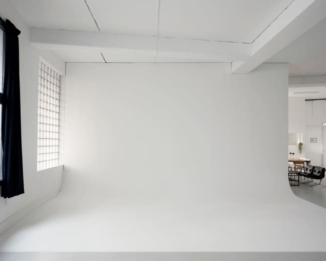 The image shows a spacious, minimalist studio with a large white backdrop, natural light flowing in from a window, and a modern seating area visible in the background.