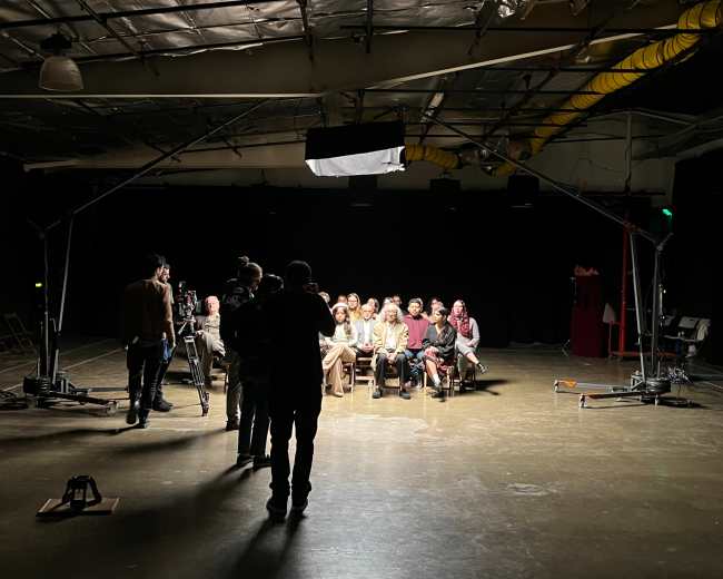 A group of seated individuals is facing a camera in a dimly lit studio, while crew members set up equipment around them.