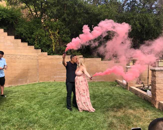 A couple stands in a grassy area, holding pink smoke grenades while surrounded by onlookers.