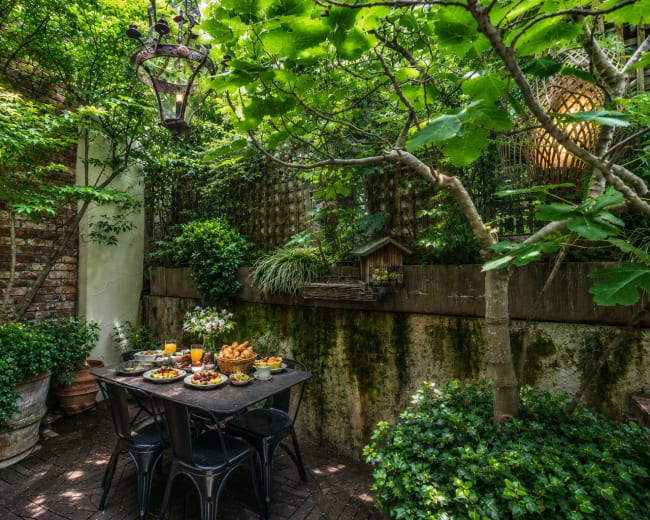 A small outdoor dining area is set with a black table, surrounded by lush greenery and potted plants against a brick wall.