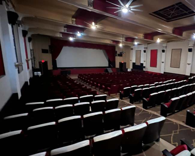 The image shows a spacious, mostly empty theater with red and white seating, a large screen, and stage lighting.