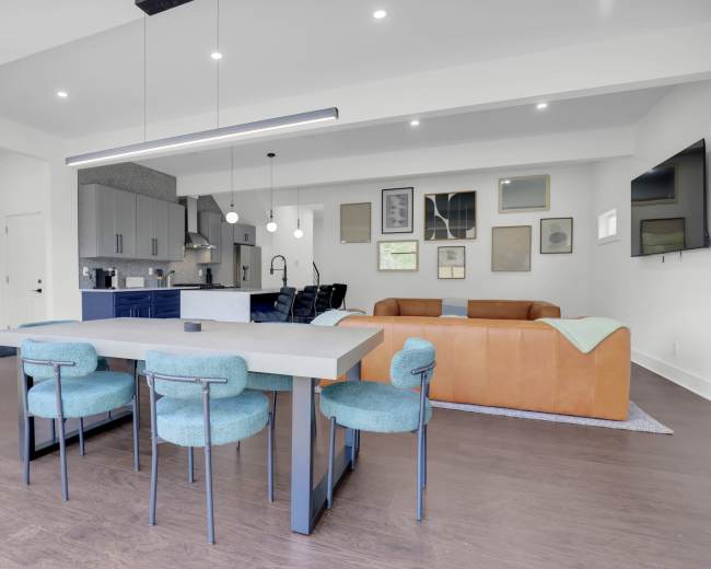 The image shows a modern open-concept kitchen and dining area featuring a rectangular table with teal chairs, a brown leather sofa, and various framed artworks on the walls.