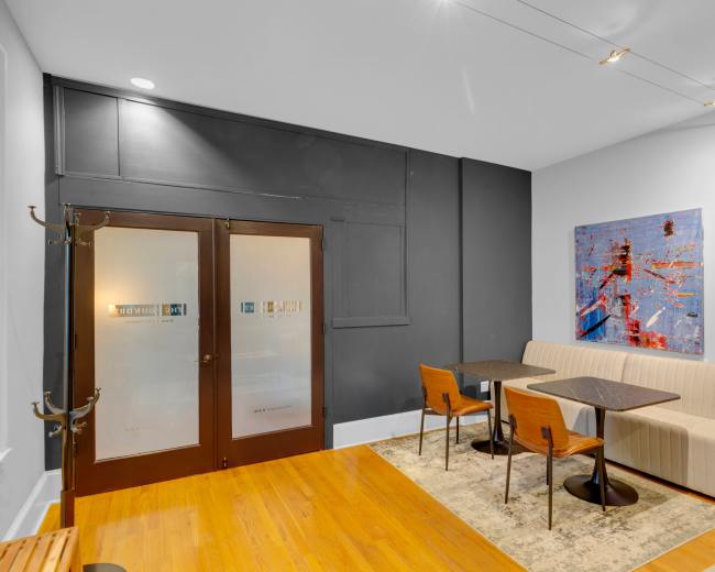 The image features a modern seating area with two tables and chairs, a bench, and a pair of glass doors leading to another room, all set against a backdrop of gray and white walls.