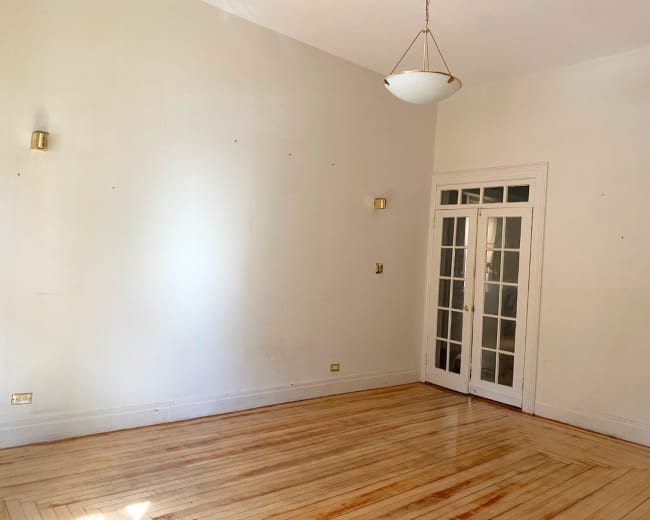 The image shows a spacious room with hardwood floors, white walls, and a set of double doors leading to another space, illuminated by a hanging light fixture.