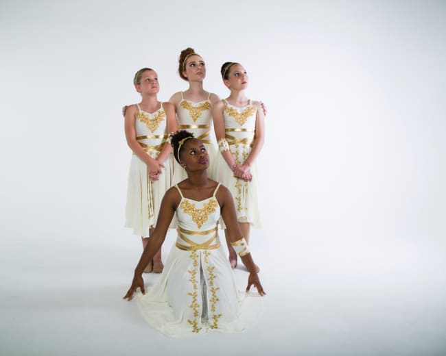 Four dancers pose in elegant white and gold costumes against a plain white background.