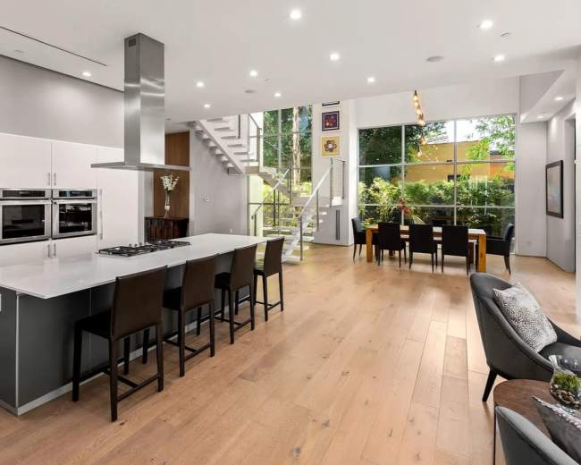 The image shows a modern open-concept kitchen and dining area with large windows allowing natural light and a view of greenery outside.