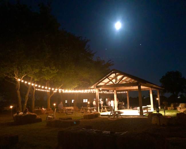 A wooden pavilion illuminated by string lights stands in a grassy area at night, with a full moon hanging overhead.