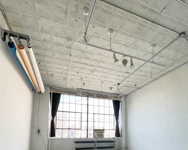 The image shows an empty, well-lit studio space with large windows, white walls, and a simple ceiling design.