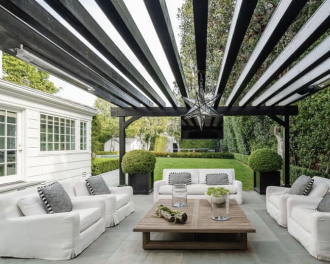 A patio features a wooden pergola with horizontal beams, surrounded by neatly trimmed hedges, and furnished with white couches and a wooden coffee table.