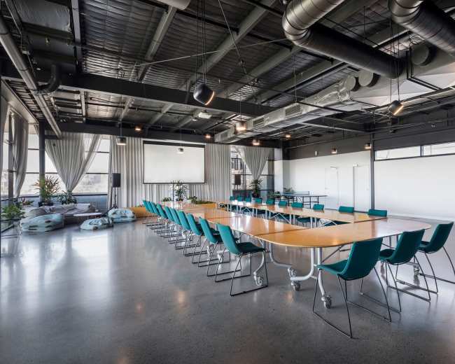 A large, modern conference room is set up with a long table and teal chairs, featuring industrial-style ceilings and large windows.