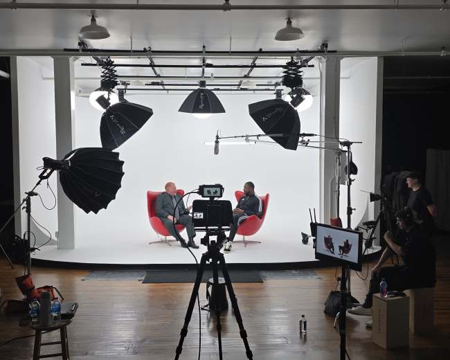 Two people sit in red chairs against a white backdrop in a well-lit studio, surrounded by camera equipment and lighting setups.