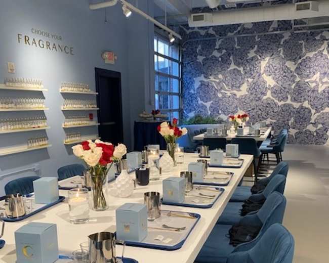 The image shows a modern, well-lit event space with a long dining table set with flower arrangements, trays, and boxes, against a decorative blue and white wall.