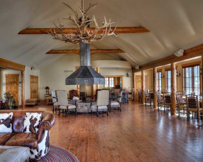 The image shows a spacious lodge interior with wooden beams, hardwood floors, and a central fireplace surrounded by seating areas.
