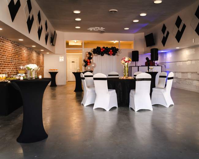 A spacious event room features black and white tablecloths arranged with chairs, decorated with floral centerpieces and a backdrop of balloons.