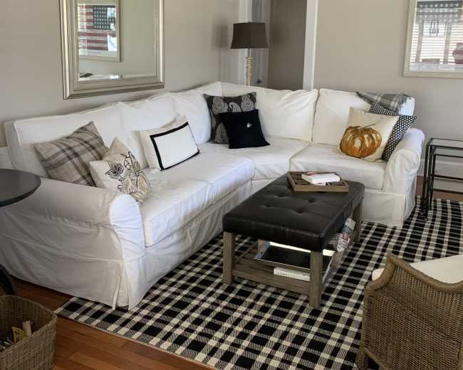 The image shows a cozy living room with a white sectional sofa adorned with various decorative pillows, a black coffee table, and a checkered area rug.