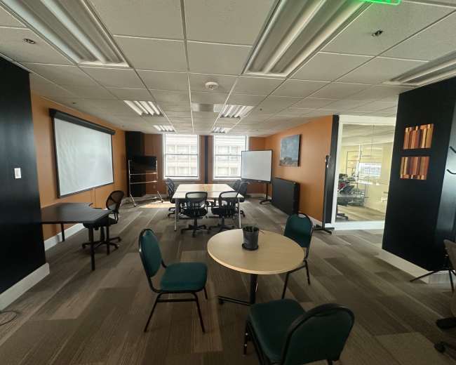 The image shows a conference room with several round tables and chairs arranged around a central area, along with a whiteboard and a projector screen on the wall.