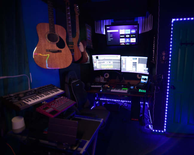 The image shows a music production room with various instruments, including guitars and keyboards, alongside dual computer monitors and colorful LED lighting.