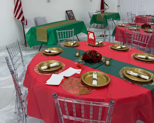The image shows a decorated event space with tables covered in red and green tablecloths, gold plates, and floral centerpieces, alongside a U.S. flag and other items on the wall.