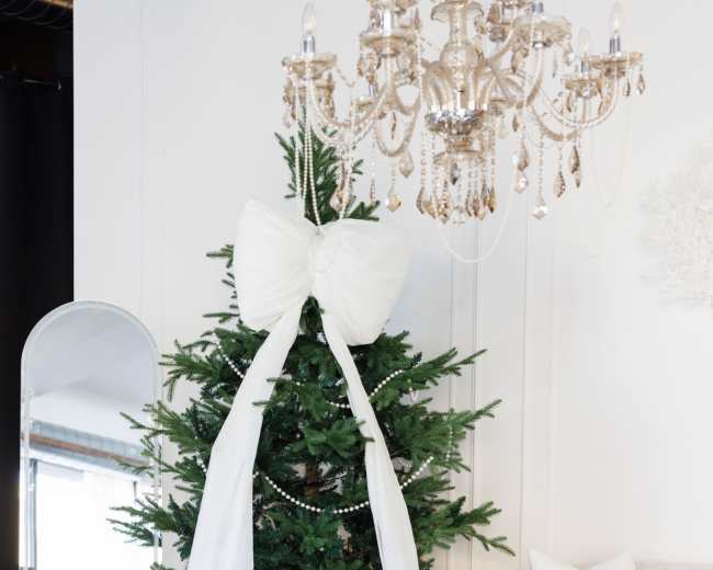 The scene features a decorated Christmas tree with a large white bow, accompanied by a chandelier, a mirror, and modern furniture in a stylish room.