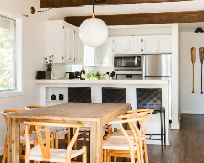 A wooden dining table with chairs is positioned in a bright kitchen area featuring white cabinets and stainless steel appliances.