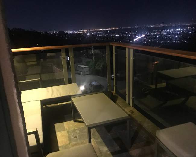 The image shows a balcony at night with modern furniture overlooking a cityscape illuminated by lights.
