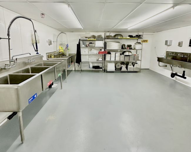 The image shows a commercial kitchen area with stainless steel sinks, shelving units, and a clean, gray floor.