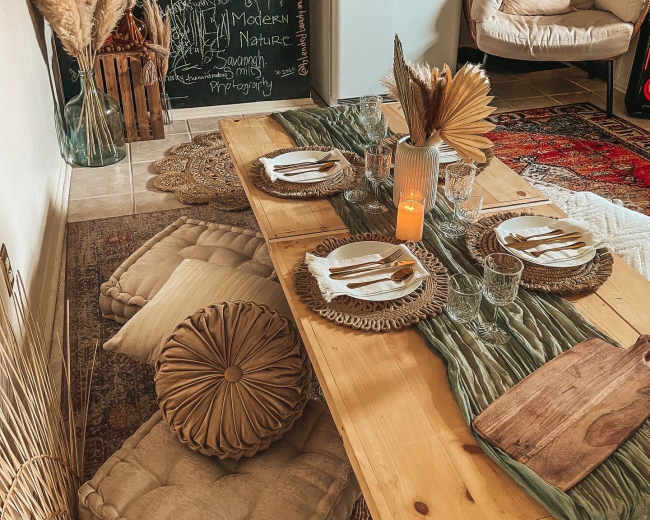 A dining space features a wooden table set with plates and glassware, surrounded by cushions, a chalkboard wall, and decorative elements like pampas grass and a glowing candle.