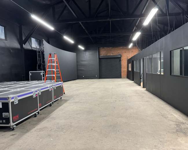 The image shows a spacious, industrial-style room with black walls, a concrete floor, a ladder against one wall, and equipment cases stacked on the floor.