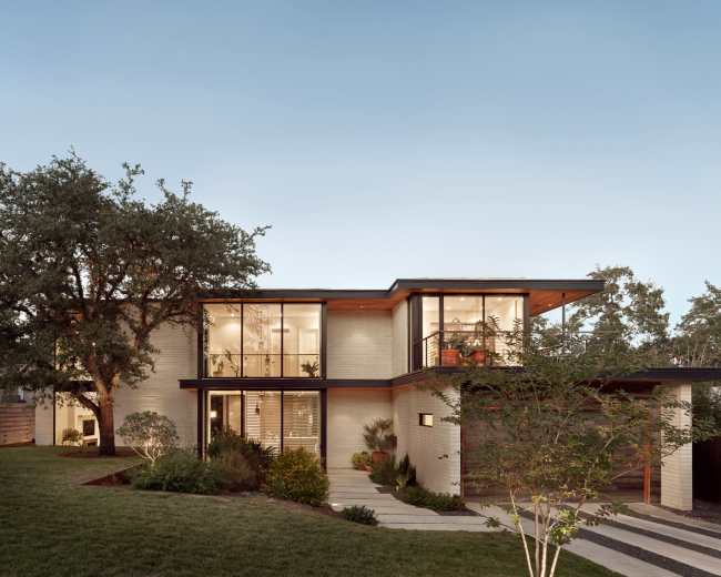 A modern two-story house features large glass windows, a wooden overhang, and landscaped greenery surrounding the property.