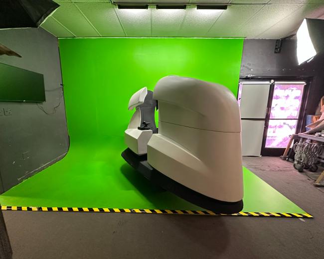 The image shows a large white piece of equipment positioned on a bright green backdrop in a studio environment.