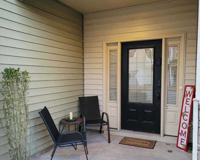 The image shows a front porch with two chairs and a small table beside a black door, featuring a "Welcome" sign.