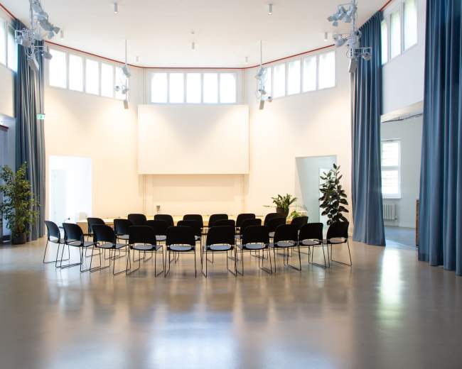 A row of black chairs is arranged in a semi-circle in a bright, minimalist room with large windows and green plants.