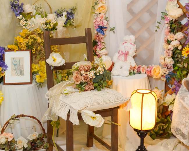 A wooden chair adorned with flowers and decorative items is set against a backdrop of draped fabric and floral arrangements, with a soft lamp illuminating the scene.