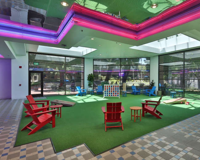 An indoor recreation area featuring colorful chairs, a large Connect Four game, and green carpeting with natural light streaming in through large windows.