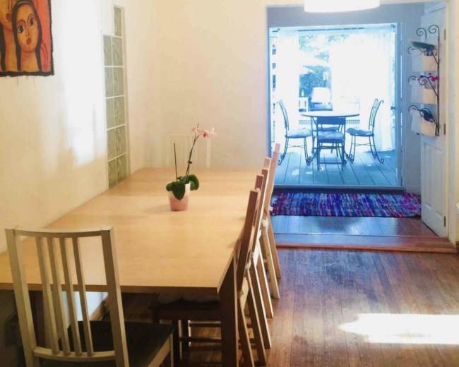 A dining area features a long wooden table with several chairs and a small potted plant, adjacent to a door leading to a deck with outdoor seating.