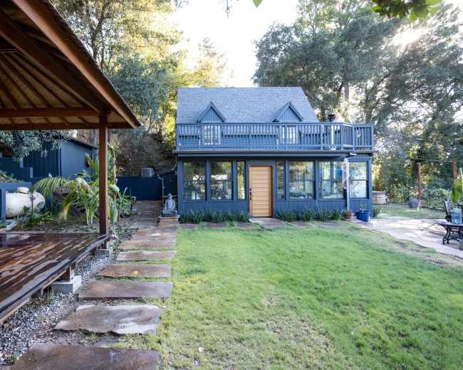 The image shows a two-story house with a peaked roof, large windows, and a wooden deck in the foreground surrounded by a neatly maintained lawn and garden.